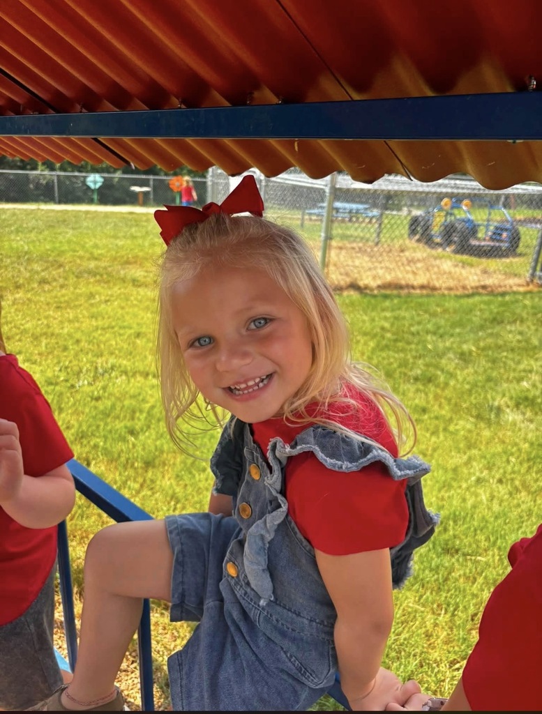 A close-up shot of a young, smiling blonde girl with blue eyes sitting outside on a playground structure. She is wearing a red shirt, denim overalls, and a large red bow in her hair.