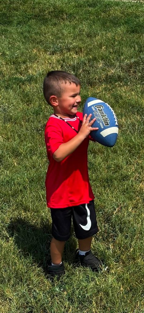 A young boy in a red t-shirt and black athletic shorts stands on a green grass field, smiling as he holds a blue and white Franklin football.
