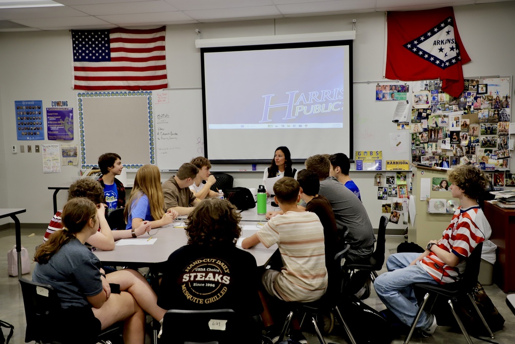 A group of high school students sitting around a classroom table, chatting and laughing together during a lunch break activity. A female instructor sits with them, holding papers and smiling at their conversation.