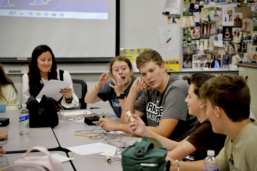 A classroom setting focusing on a group of high school students at a table. A boy in a grey shirt leans his head on his hand while holding a quiz buzzer, listening intently. Next to him, a girl gestures with her hands while speaking excitedly, and an instructor sits in the background.