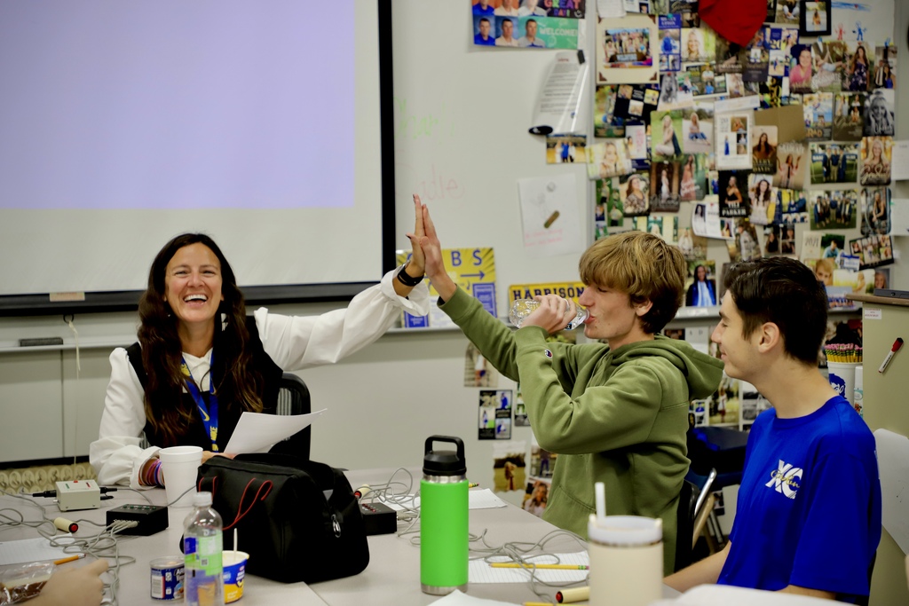 A close-up of a smiling female instructor giving a high-five across a classroom table to a male student in a green hoodie who is drinking water. Another student in a blue t-shirt looks on. A colorful bulletin board filled with photos is visible in the background.