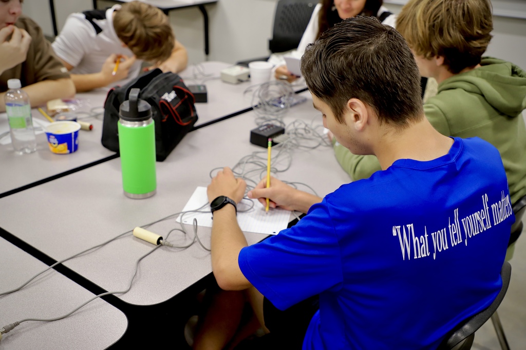 Over-the-shoulder view of a student wearing a blue shirt that reads "What you tell yourself matters" on the back. He is sitting at a classroom table filled with tangled buzzer wires, writing on a piece of notebook paper with a pencil.
