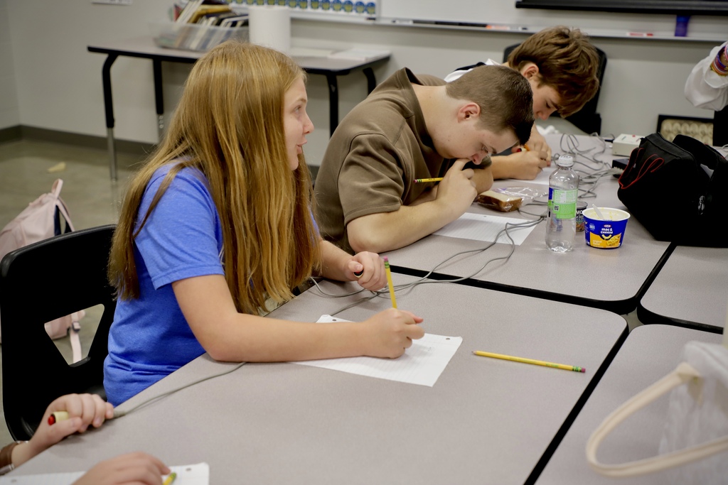 A close-up profile shot of a high school girl with long blonde hair wearing a blue shirt, sitting at a classroom table and writing on a piece of paper with a pencil. Next to her, a boy in a brown shirt is also focused on writing.
