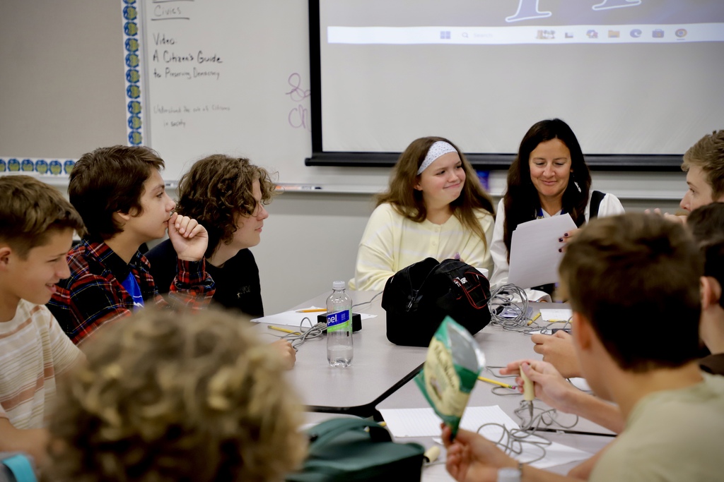 A group of high school students sitting around a classroom table, chatting and laughing together during a lunch break activity. A female instructor sits with them, holding papers and smiling at their conversation.