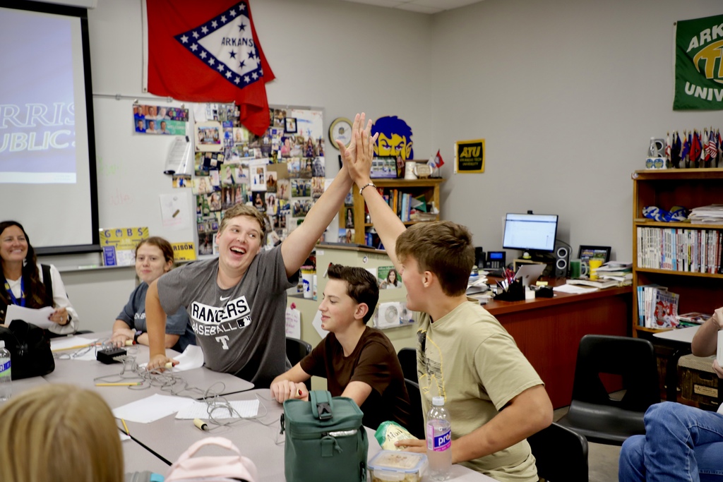 A joyful classroom scene where two high school boys are mid-air giving each other an enthusiastic high-five across a table. One boy wears a grey "Rangers Baseball" shirt and has a wide smile, while a female instructor and other students watch happily.