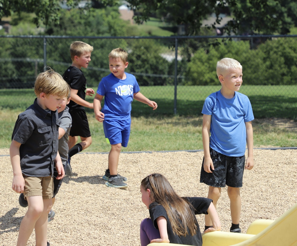 A group of elementary school boys run and play happily on a pebble-filled playground area on a sunny day.