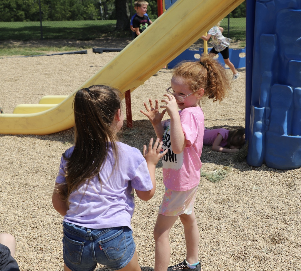Two young elementary school girls smile and talk playfully to each other next to a yellow playground slide during recess.