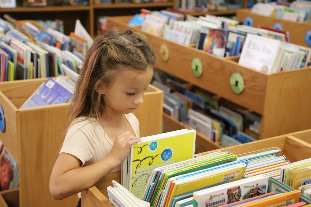 A young elementary-aged girl with her hair partially braided stands in a school library, carefully looking through a bin of colorful picture books and resting her hand on a yellow book.