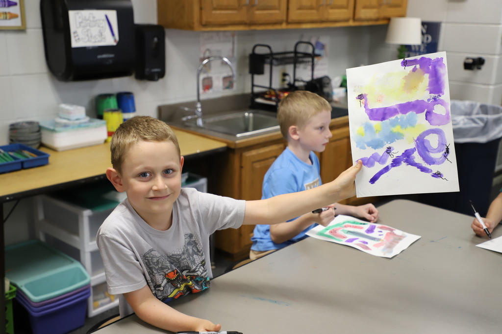 A young elementary school boy smiles at the camera as he proudly holds up his finished abstract purple and blue watercolor painting in an art classroom.