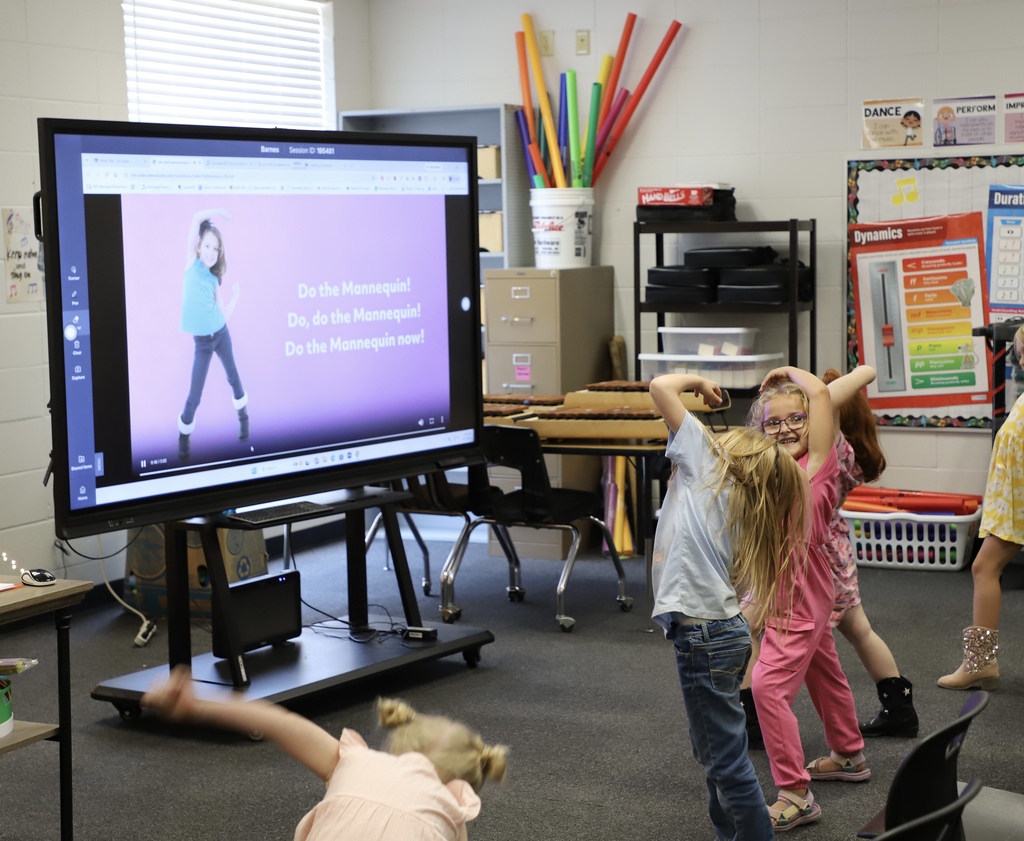 Young elementary school girls joyfully dance and mimic a "Do the Mannequin" dance video playing on a large interactive smart screen at the front of a classroom.
