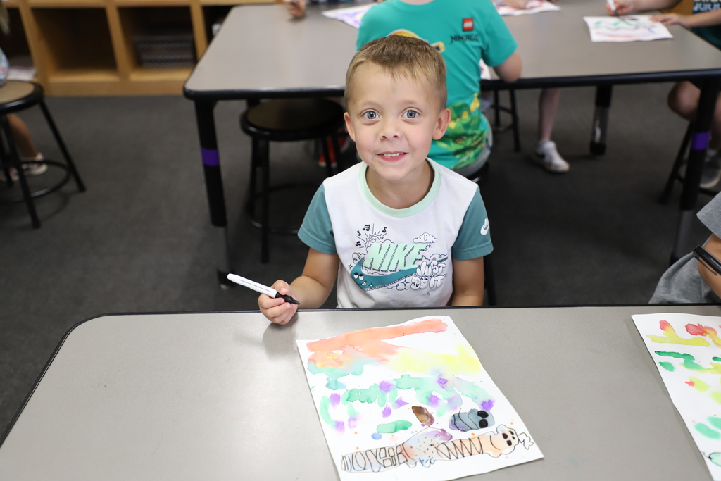A smiling young boy in a white Nike shirt sits at a school desk, proudly holding a black marker over his colorful watercolor painting.