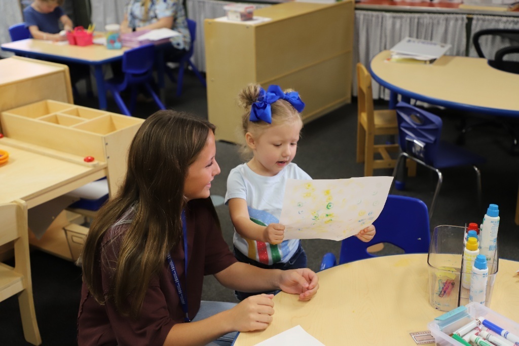 A teenage girl with long brown hair smiles warmly at a young preschool girl with a large blue hair bow. The child is proudly holding up a colorful, stamped piece of artwork to show her at a low classroom table.