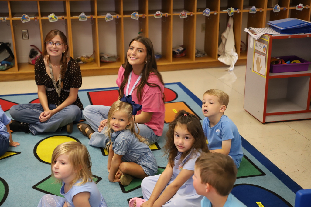 Two teenage girls and several young preschool children sit together on a colorful geometric-patterned rug inside a classroom. One teenage girl wears a black polka dot shirt, and the other wears a pink shirt. They are looking toward the camera while the children face forward, listening attentively.