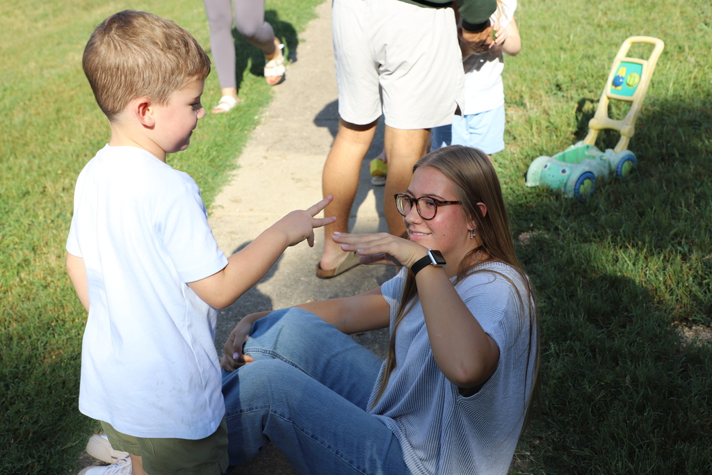 A teenage girl wearing glasses and a gray shirt sits on the grass outdoors, smiling as she plays a game of rock-paper-scissors with a young preschool boy standing in front of her.