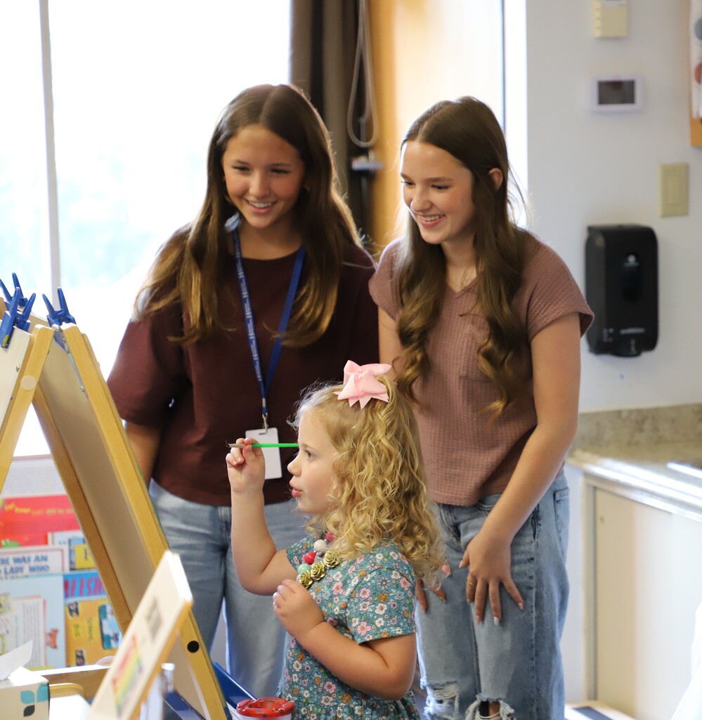 Two teenage girls with long brown hair smile as they watch a young preschool girl with a pink bow in her hair practice painting at a wooden easel inside a brightly lit classroom sink area.