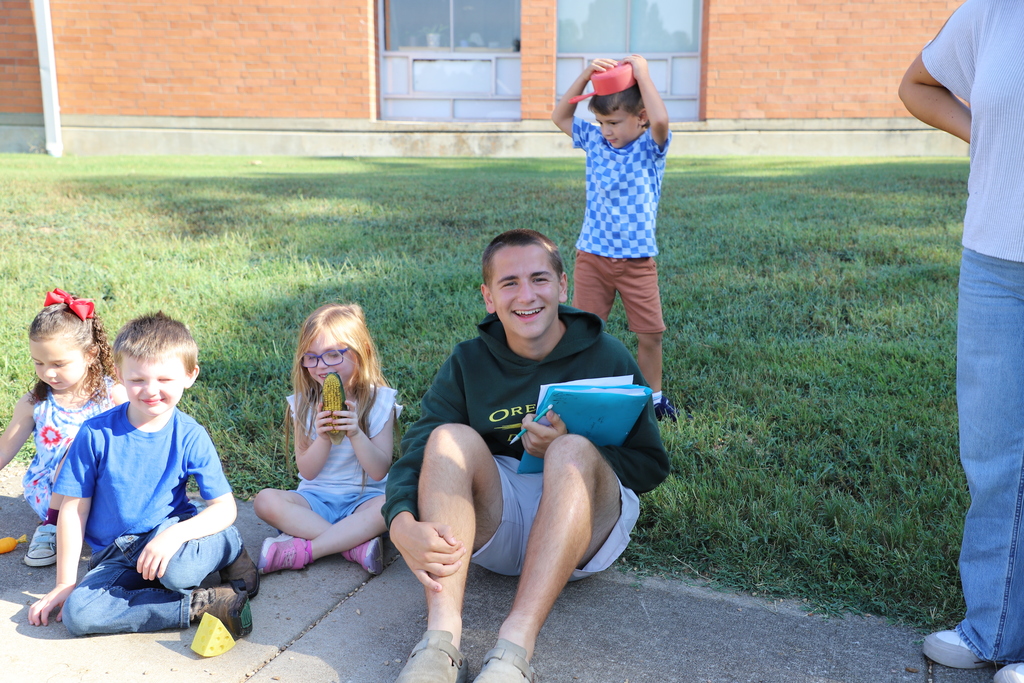 A smiling teenage boy wearing a green Harrison High School sweatshirt sits on a concrete sidewalk outdoors next to three preschool children. He is holding a blue folder and a pen. In the background, a young boy in a blue checkered shirt balances a small red toy bowl on his head in a grassy field.