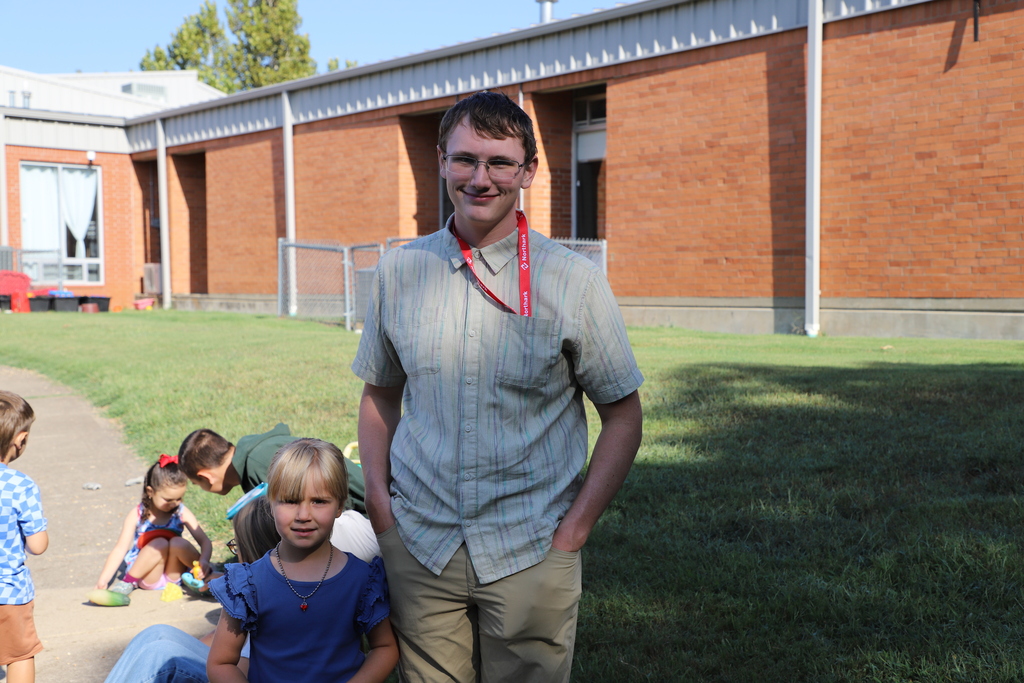 A teenage boy wearing glasses, a button-down shirt, and a red Northark lanyard stands outside on the grass smiling at the camera, next to a young preschool girl in a blue shirt. Other children play in the background near a brick school building.