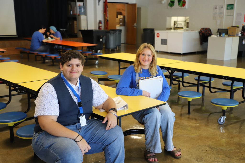A teenage boy in a blue vest over a patterned shirt and a teenage girl in a blue sweatshirt and denim overalls sit at a yellow cafeteria table holding school binders and smiling at the camera.