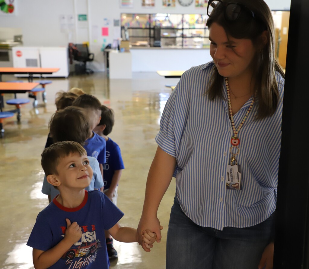 teacher or caregiver in a blue-and-white striped shirt leads a line of young children through what looks like a school cafeteria or hallway.