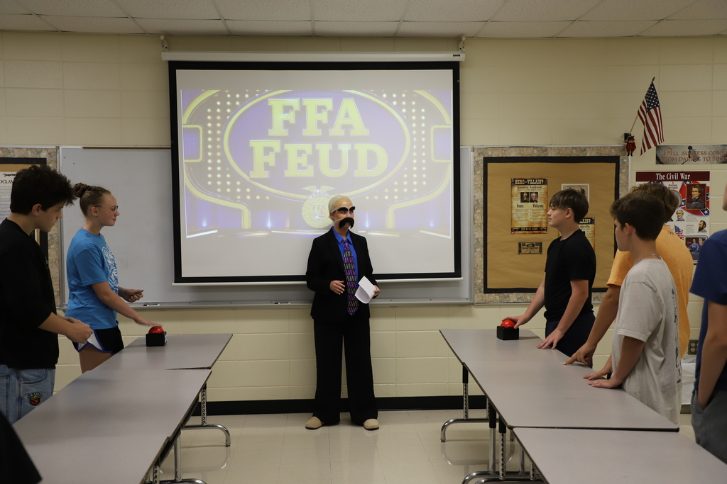 Middle school students standing at tables with red buzzers on either side of their teacher, who is dressed as game show host Steve Harvey in front of a projection reading "FFA Feud."