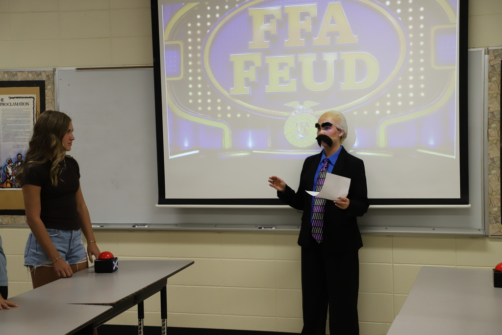An agriculture teacher dressed up as Steve Harvey holding notes and speaking to a student standing behind a table with a red game buzzer during an "FFA Feud" classroom activity.