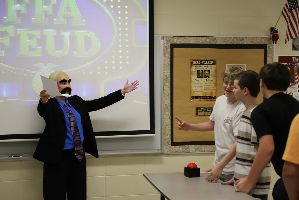 An agriculture teacher dressed in a suit, mustache, and bald cap like Steve Harvey holding papers with arms wide open, hosting an "FFA Feud" game for 8th-grade middle school students.