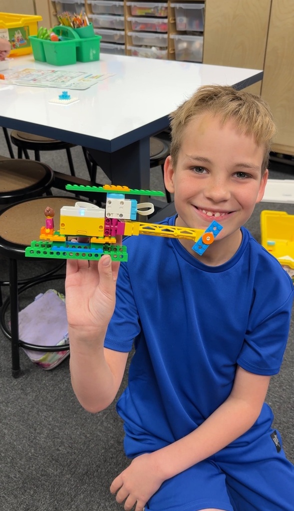 A young boy smiling while holding up a custom helicopter made from colorful plastic building bricks and motor components inside a classroom.