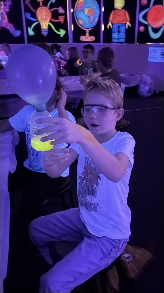 A young boy wearing safety glasses in a dark room illuminated by blacklight, holding up a plastic bottle filled with glowing yellow liquid with a blue balloon attached to the top.