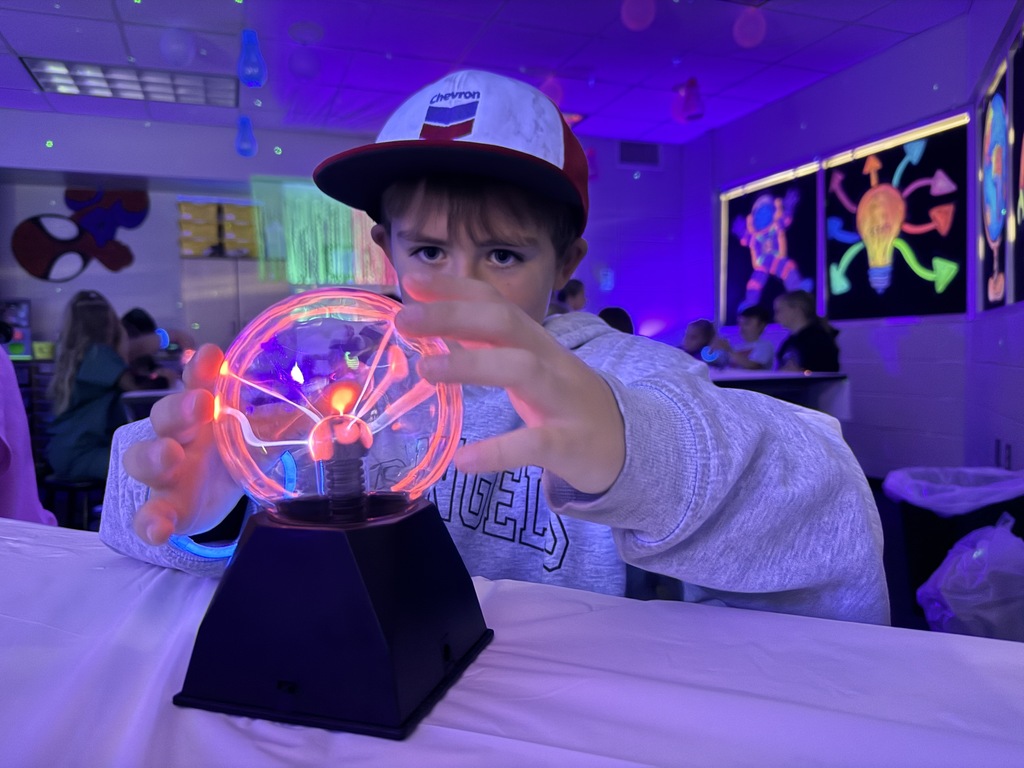 A young boy wearing a Chevron trucker hat placing his hands near a glowing plasma ball on a table in a dark, UV-lit classroom setting.