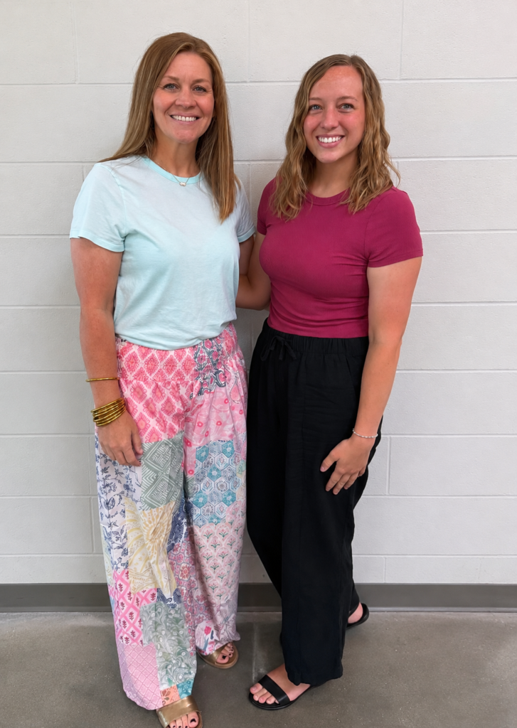A full-length photo of two smiling women standing side-by-side in front of a white brick wall. The woman on the left wears a light blue t-shirt and colorful patchwork wide-leg trousers, while the woman on the right wears a dark pink t-shirt and black pants.