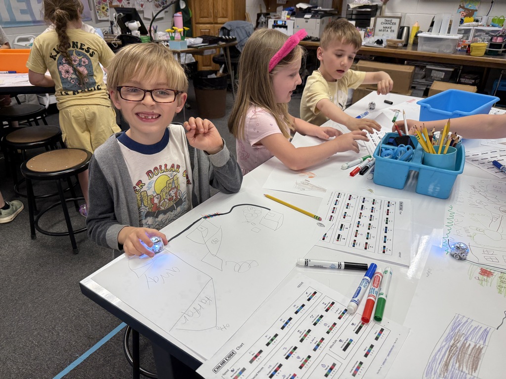 A young boy in a grey shirt and glasses smiling at a classroom table while drawing black and colorful track lines on paper to guide a small floor robot. Other elementary students work nearby at the same table.