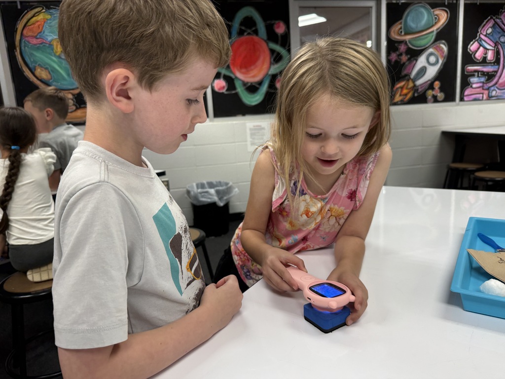Two young children leaning over a white table in a classroom, using a digital handheld device to examine an object up close.