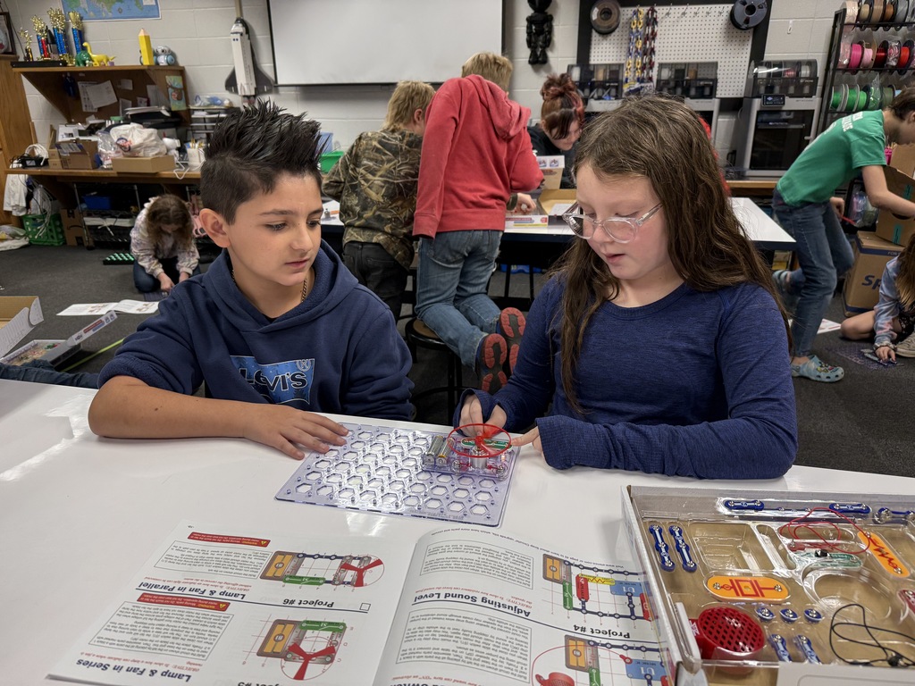 A young boy and girl sitting together at a table in a classroom, actively building an electronic circuit project on a clear grid using a Snap Circuits set with a fan and lamp module.