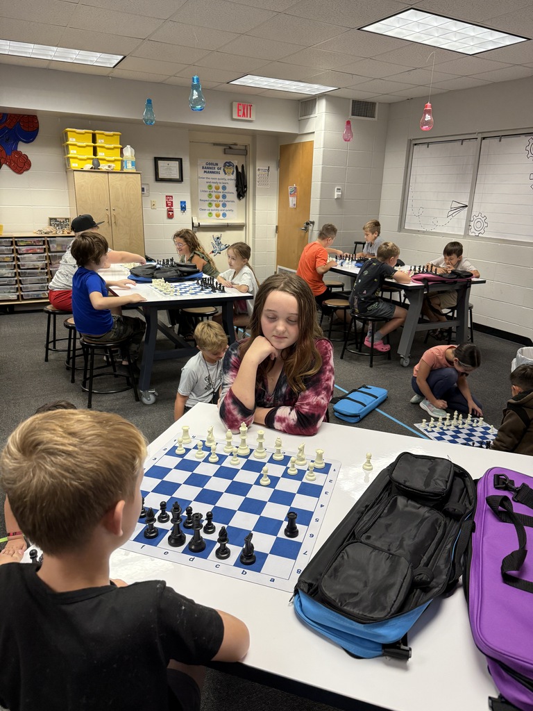 A wide shot of an elementary classroom filled with students sitting at group tables playing chess on blue and white vinyl chess boards. In the foreground, a girl wearing a tie-dye shirt concentrates on her chess pieces.
