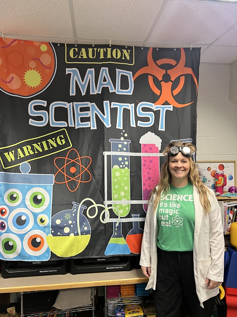 A teacher wearing a white lab coat, green "Science: it's like magic but real" t-shirt, and safety goggles over her forehead smiling in front of a black banner with "Caution Mad Scientist" written above colorful beaker illustrations.