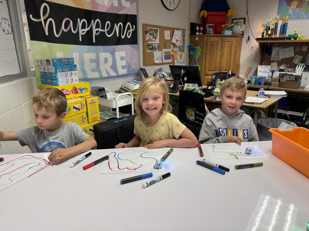 Three young elementary students sitting side-by-side at a white classroom table, smiling while using markers to draw colorful paths on large paper sheets to direct small blue floor robots. A banner reading "Happens Here" hangs on the wall behind them.