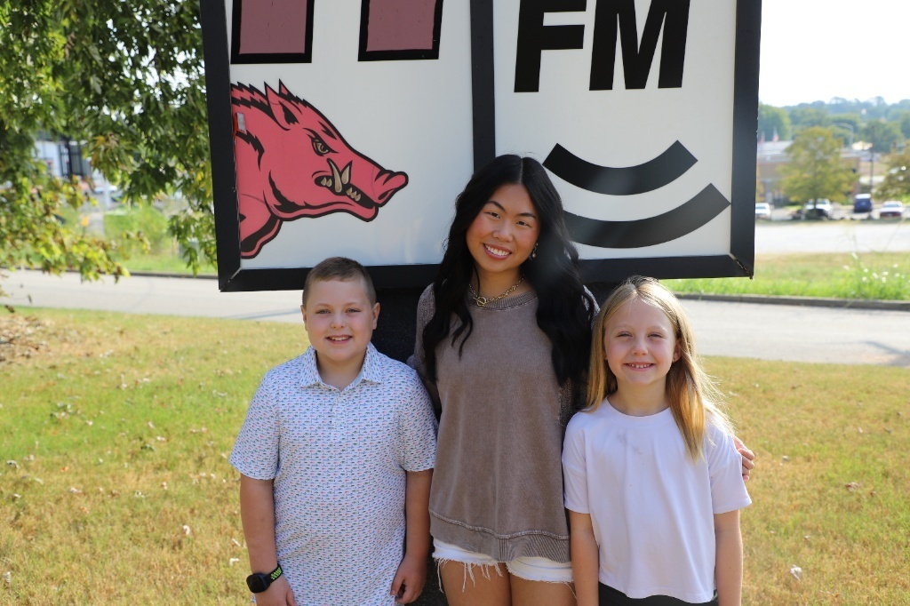 Medium group shot of students Oliver Mathis, Stevie Jones, and Olivia Reeves posing together in front of the ESPN 104.3 FM sign.