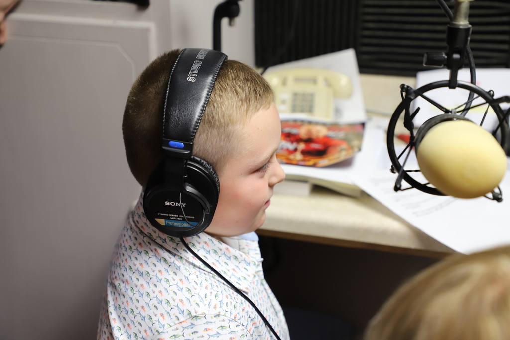 A young boy with short hair wearing headphones and sitting in front of a radio studio microphone.