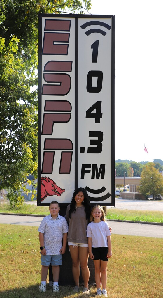 Full-length photo of students Oliver Mathis, Stevie Jones, and Olivia Reeves standing outside in front of the tall ESPN 104.3 FM outdoor sign.