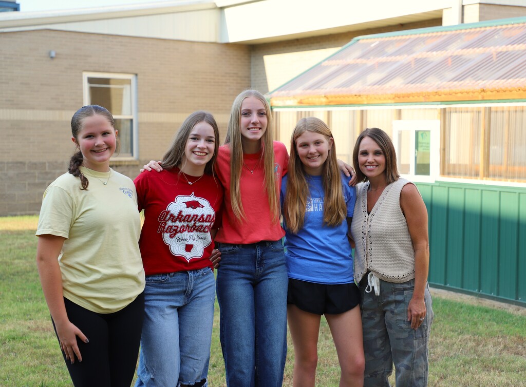 Four middle school girls stand alongside their female teacher outdoors near a school greenhouse, smiling with arms around each other.