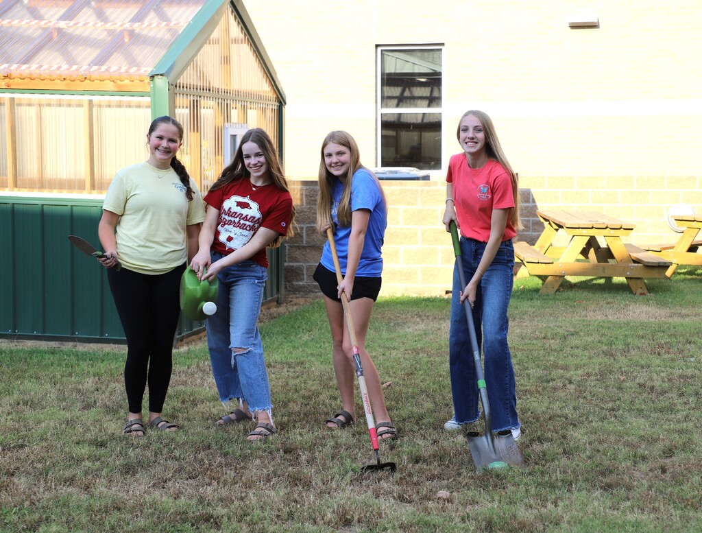 Four middle school girls pose outside on a lawn holding gardening tools, including a trowel, a green watering can, a hoe, and a shovel. A school greenhouse and picnic tables stand in the background.