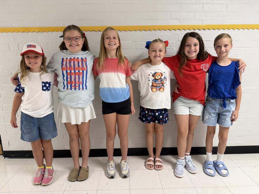 Six young girls standing shoulder-to-shoulder against a white brick wall in a school hallway wearing patriotic clothing.