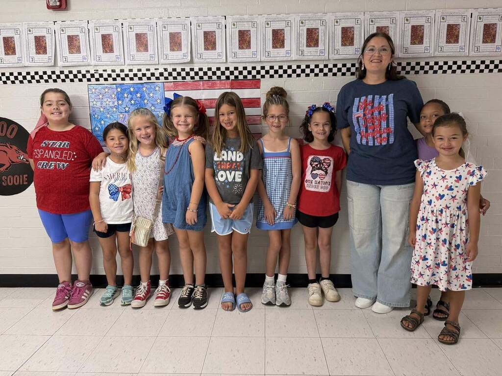 A teacher standing with a line of elementary school students dressed in red, white, and blue shirts in a classroom hallway.