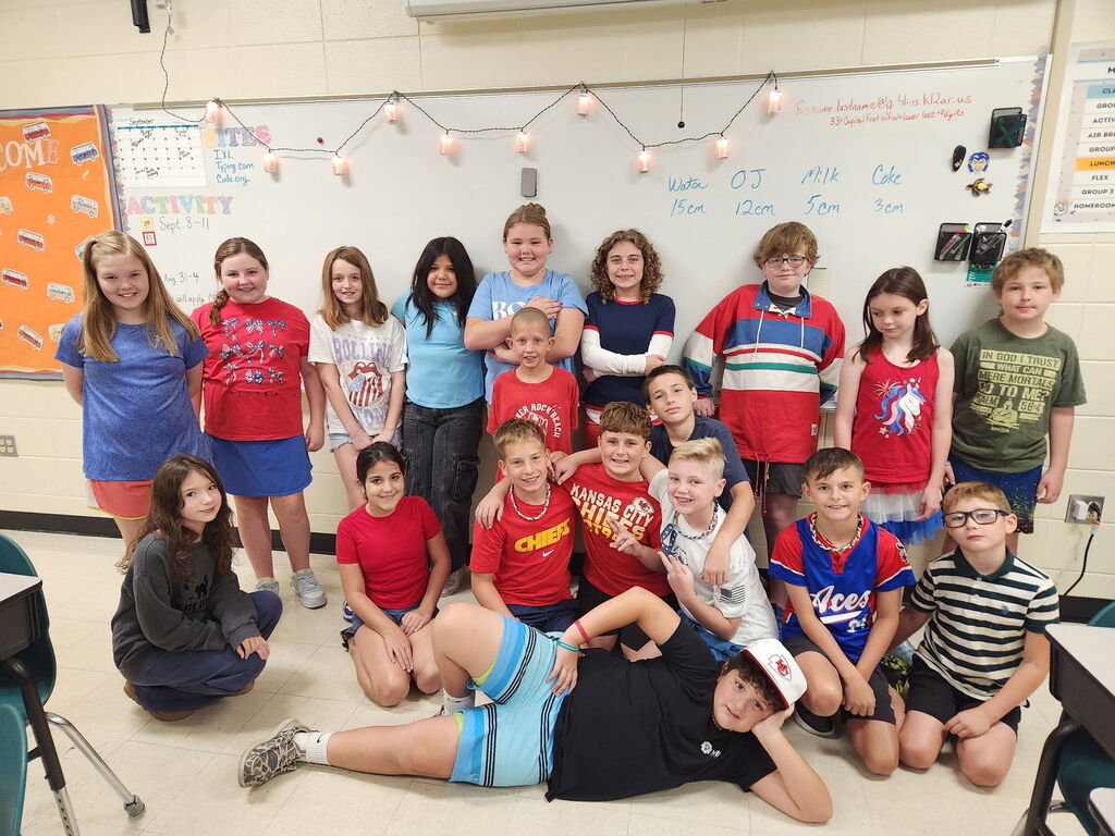 A group of elementary school students in red, white, and blue clothing posing together in a classroom in front of a whiteboard, with one student playfully reclining across the floor in front.