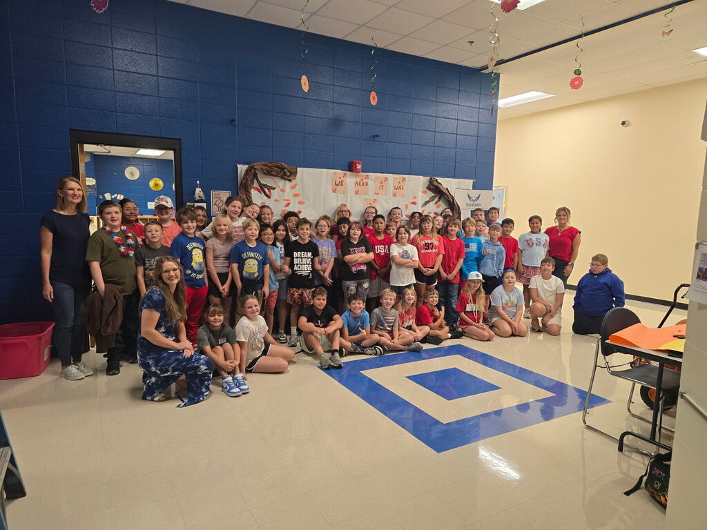A large group of elementary students and teachers wearing patriotic attire posing together in a multipurpose room with a blue square floor pattern and fall bulletin board display.