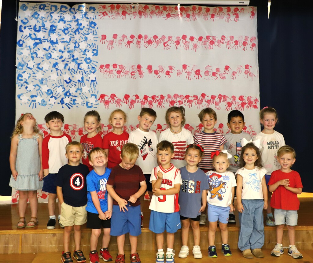 A group of elementary students standing on a stage in front of a giant paper American flag created using red and blue handprints.