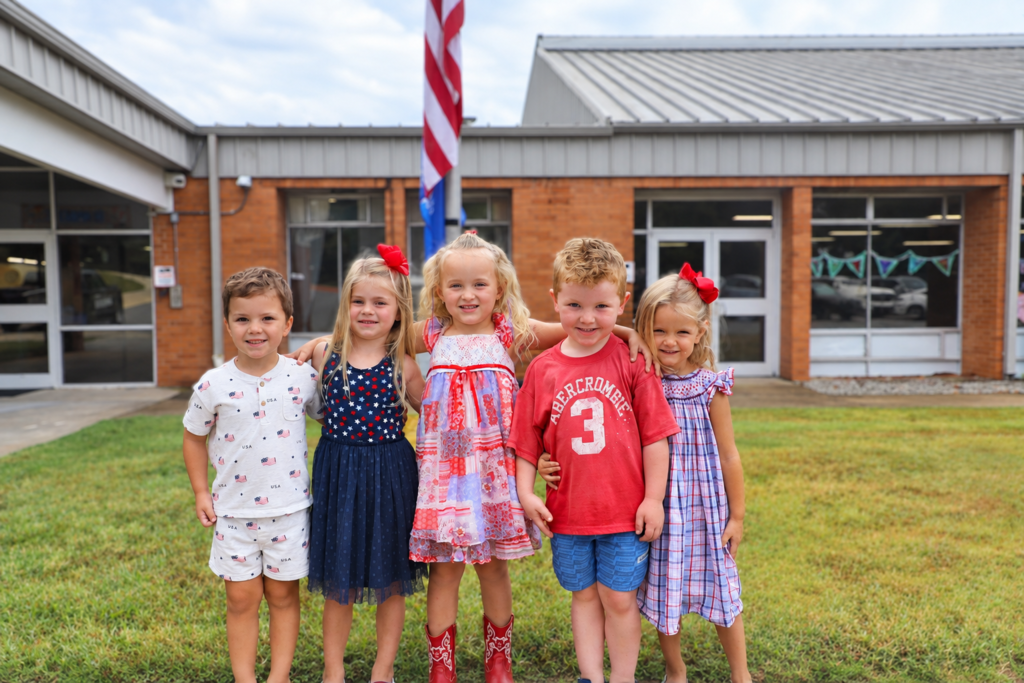 Five young school children standing close together outdoors in front of an elementary school building with an American flag flying in the background. IMG_0205.jpg