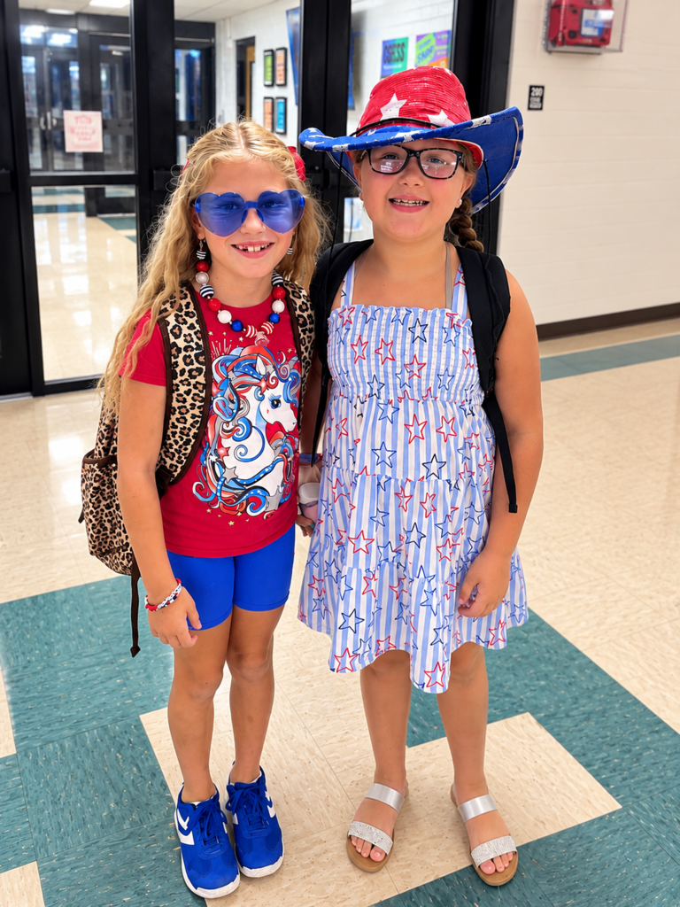 Two young girls wearing patriotic outfits, red-white-and-blue cowboy hat, and heart-shaped sunglasses posing inside a school corridor.