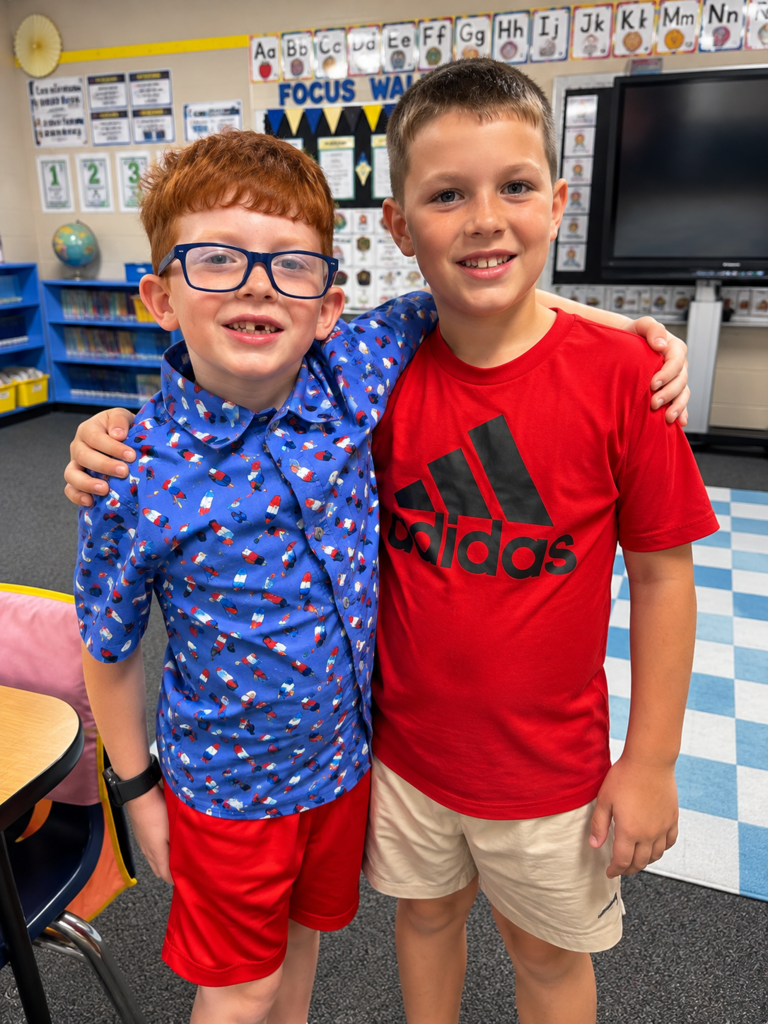 Two young elementary boys smiling and hugging with their arms around each other inside a classroom, dressed in red and blue tops.
