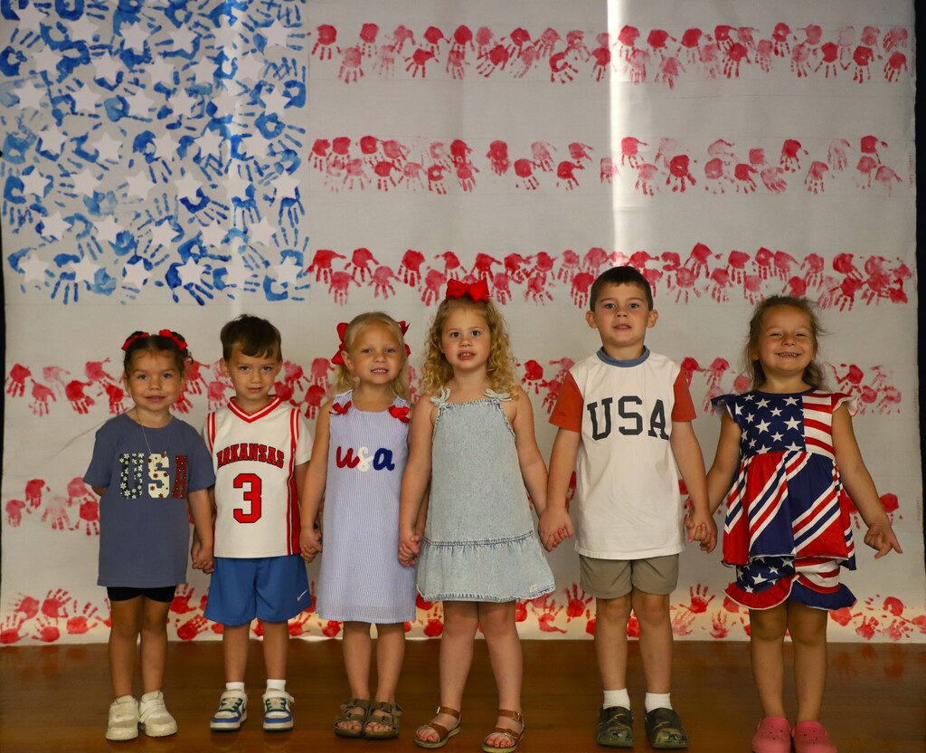 Six young elementary students standing side-by-side in patriotic attire, holding hands in front of a large handprint-painted American flag poster.